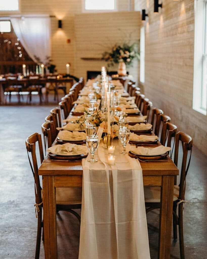 long table with wood chairs, plates, and tall candle centerpieces inside white barn